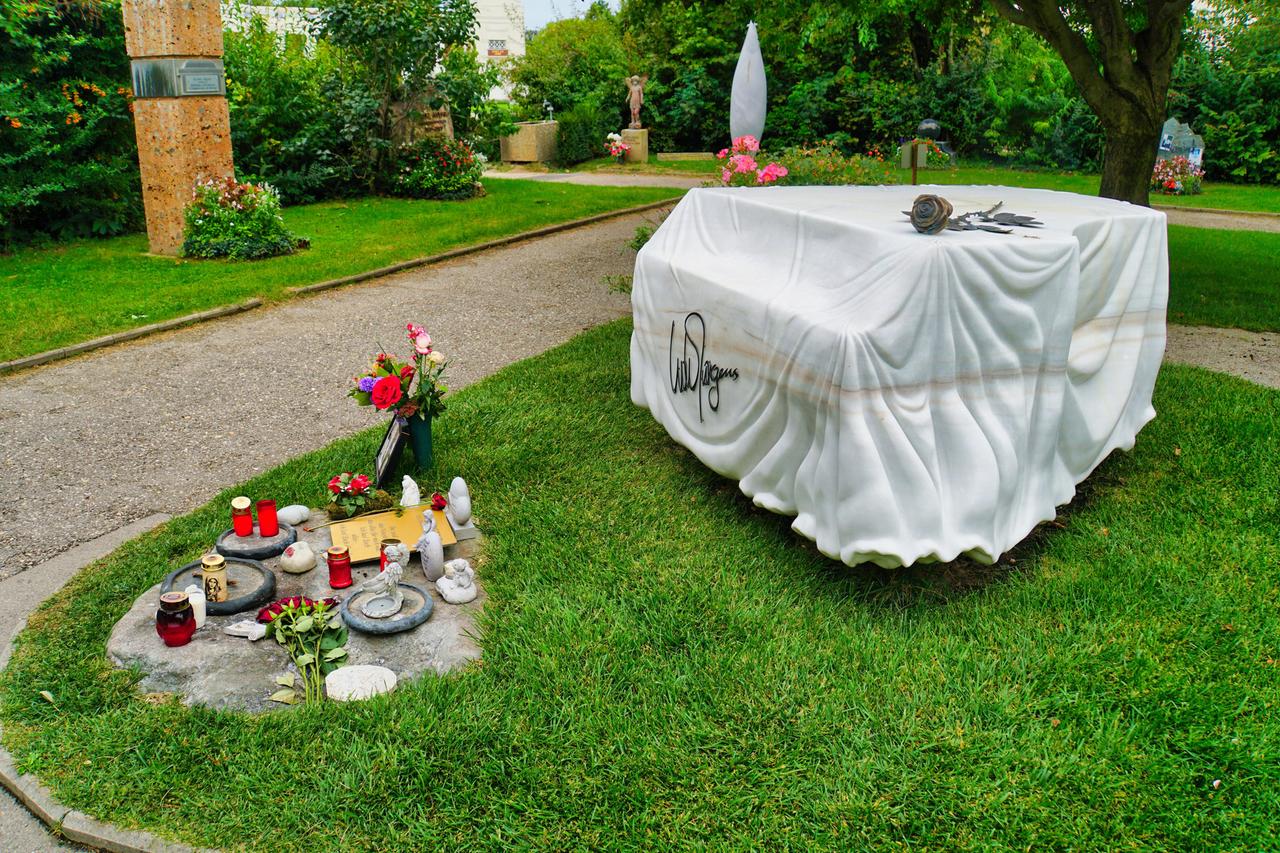 Blick auf das Grab von Udo Jürgens auf dem Zentralfriedhof Wien. Zu sehen ist ein weißer Marmorstein, der einen mit Tuch abgedeckten Flügel (das Musikinstrument) darstellen soll. Darauf liegt eine Rose. 