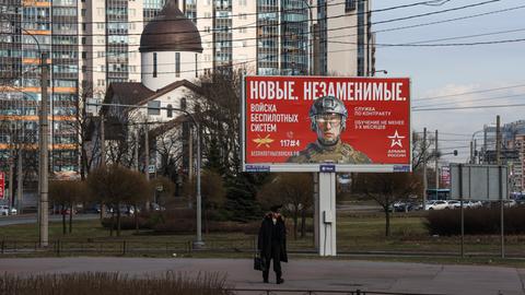Ein Kadett geht an einer Außenwerbetafel in St.Petersburg vorbei, auf der unter anderem „Neue. Unersetzliche“ steht sowie Informationen zur Rekrutierung für die Einheiten für unbemannte Systeme der russischen Streitkräfte. 

