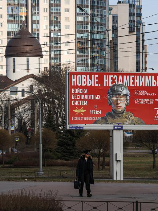 Ein Kadett geht an einer Außenwerbetafel in St.Petersburg vorbei, auf der unter anderem „Neue. Unersetzliche“ steht sowie Informationen zur Rekrutierung für die Einheiten für unbemannte Systeme der russischen Streitkräfte. 
