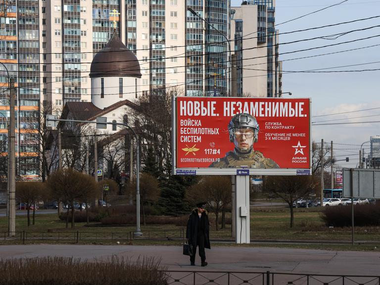 Ein Kadett geht an einer Außenwerbetafel in St.Petersburg vorbei, auf der unter anderem „Neue. Unersetzliche“ steht sowie Informationen zur Rekrutierung für die Einheiten für unbemannte Systeme der russischen Streitkräfte. 
