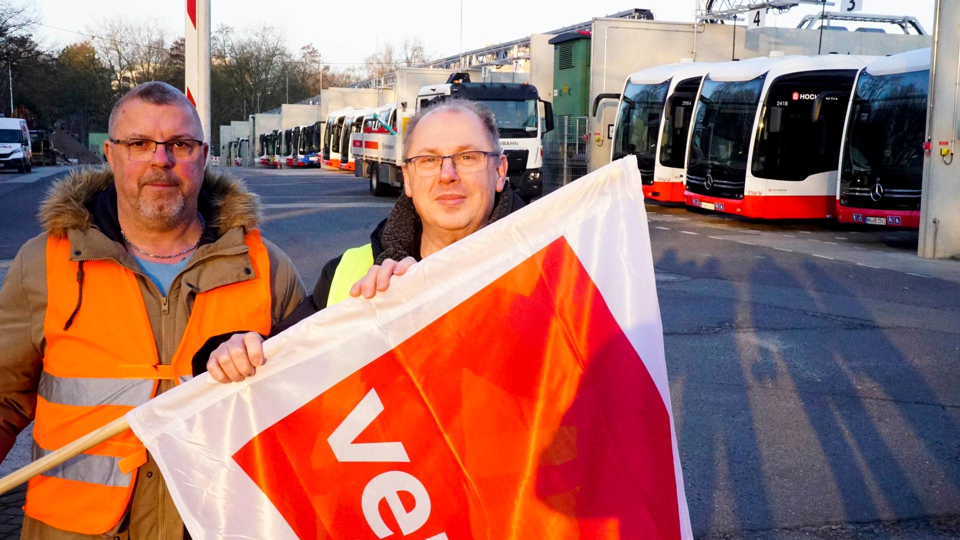 Zwei Männer stehen vor einer Reihe von Bussen. Sie haben eine Fahne in der Hand. Auf der Fahne steht verdi. verdi ist eine Gewerkschaft. 