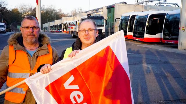 Zwei Männer stehen vor einer Reihe von Bussen. Sie haben eine Fahne in der Hand. Auf der Fahne steht verdi. verdi ist eine Gewerkschaft. 
