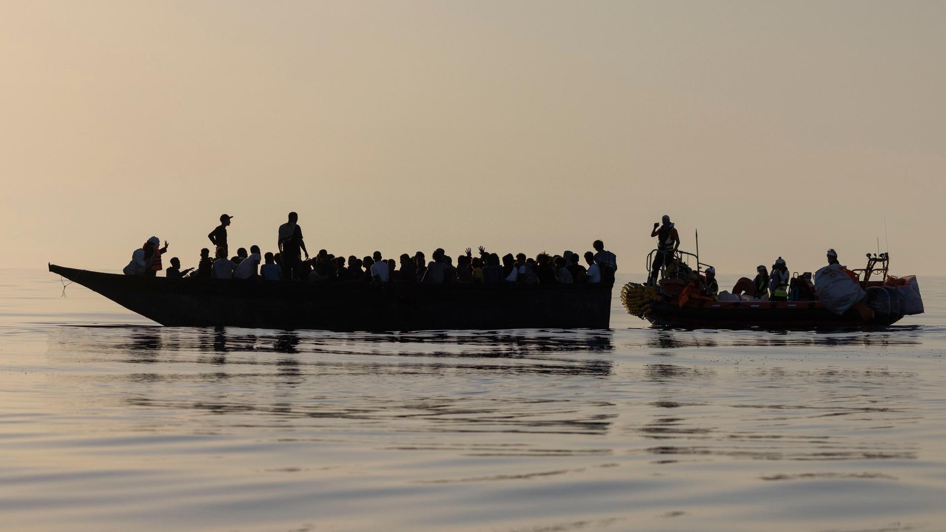 Migranten in einem Boot im Mittelmeer werden von einem anderen Boot gerettet.