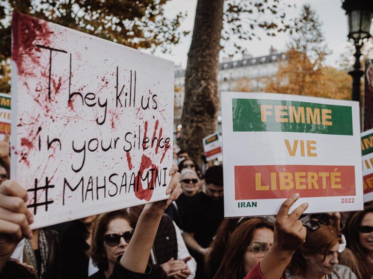 Demonstrierende halten Plakate mit der Aufschrift "They kill us in your silence" und "Femme Vie, Lieberté"