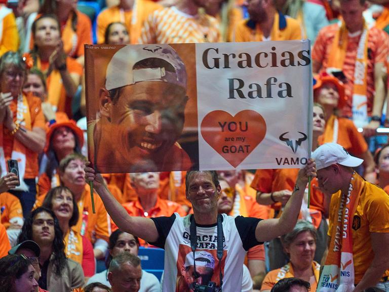 Zu sehen sind Fans, die ein Banner hochhalten, auf dem ein Foto von Rafael Nadal gedruckt ist und "Gracias Rafa" steht.