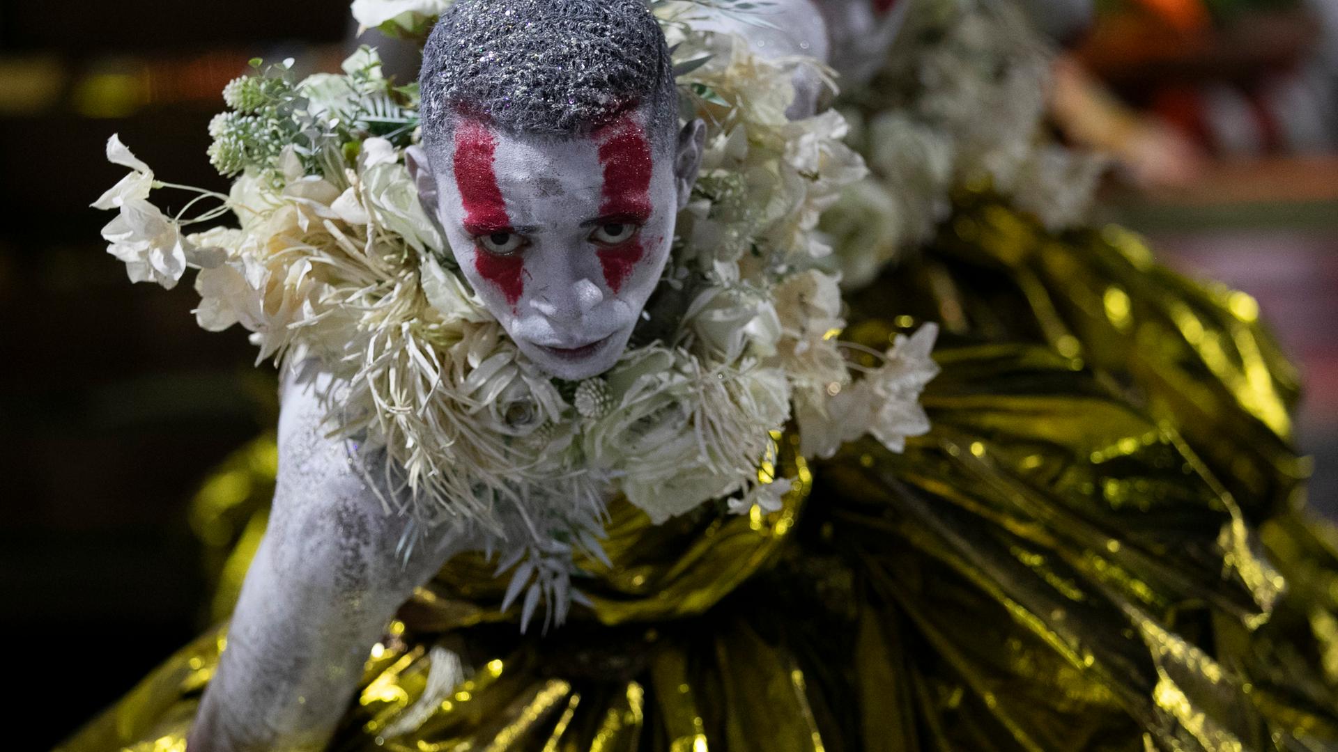 Ein Mitglied der Samba-Schule "Beija-Flor" tanzt. Er trägt gold glänzende Kleidung und Blumen. Er ist weiß geschminkt mit roten Streifen.