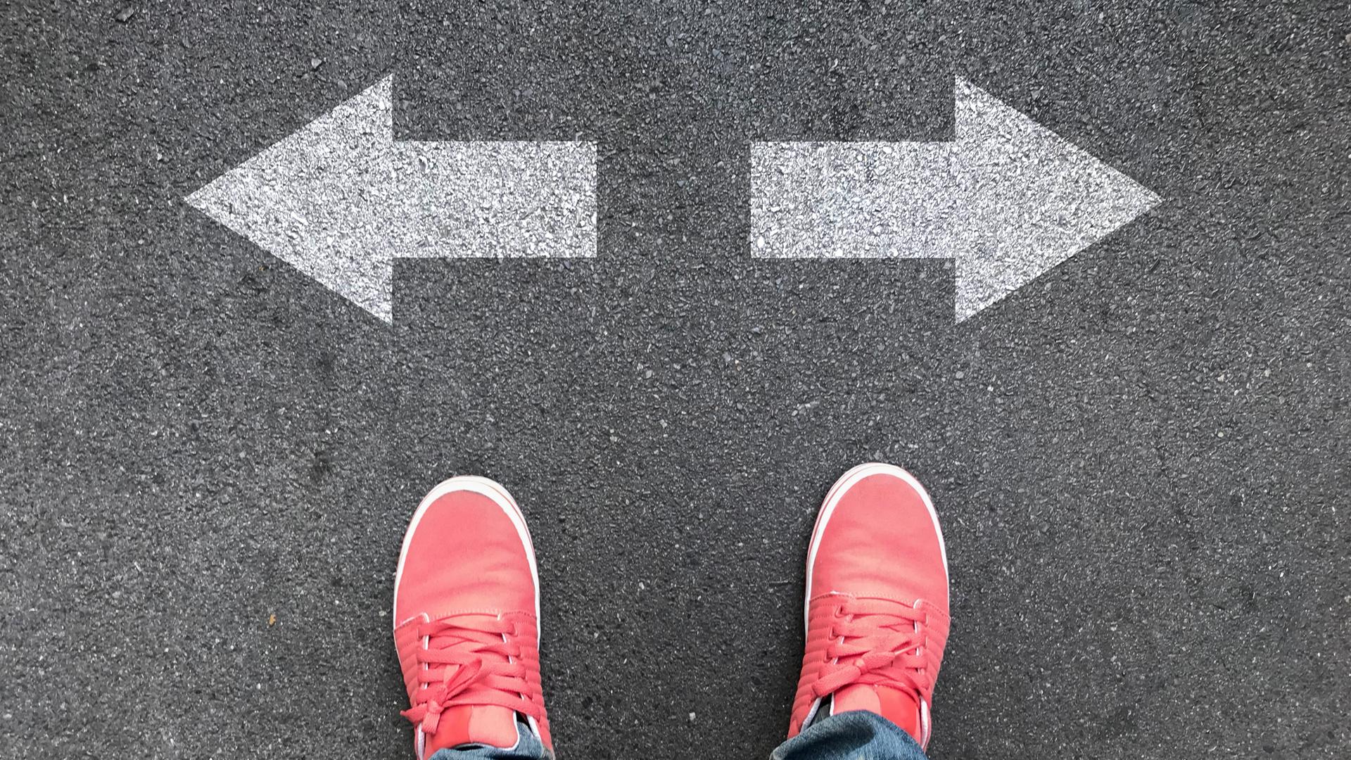 Blick von oben auf ein Paar pinke Schuhe auf Asphalt, die vor einem Pfeil, der nach links und einem Pfeil, der nach rechts zeigt, stehen.