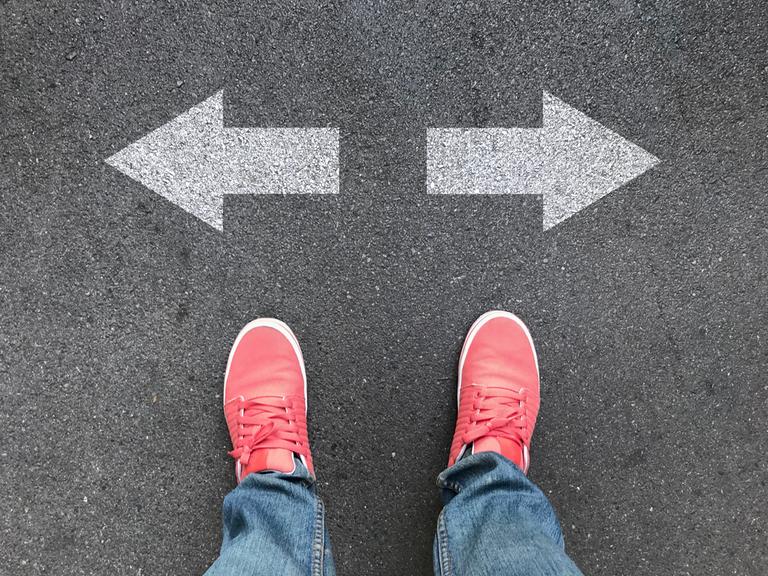 Blick von oben auf ein Paar pinke Schuhe auf Asphalt, die vor einem Pfeil, der nach links und einem Pfeil, der nach rechts zeigt, stehen.