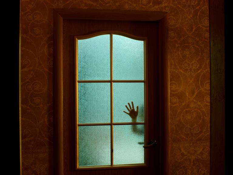 Eine Kinderhand an einer geschlossenen Glastür, in gruseliger Atmosphäre.