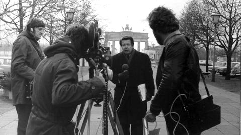 Der Journalist Lothar. Ein Kamerateam und ein Reporter mit Mikrofon in der Hand stehen vor dem Brandenburger Tor.