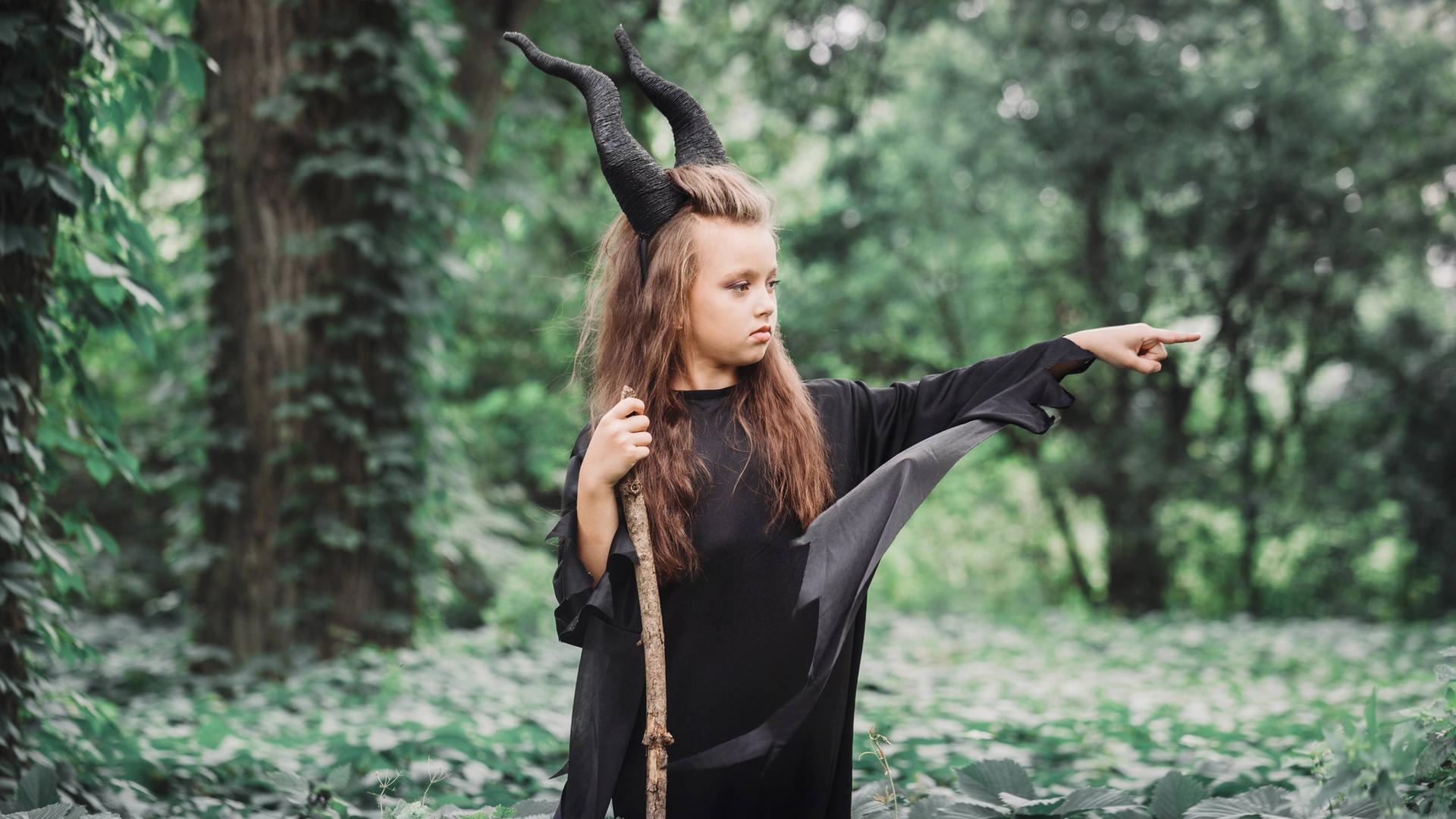 Ein Mädchen steht mit schwarzen Hörnern, Hexenkleid und einem Stock im Wald und streckt wütend seinen Finger aus.