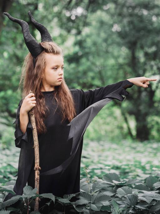 Ein Mädchen steht mit schwarzen Hörnern, Hexenkleid und einem Stock im Wald und streckt wütend seinen Finger aus.