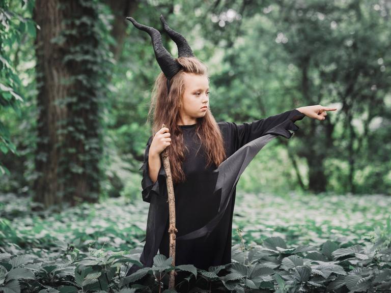 Ein Mädchen steht mit schwarzen Hörnern, Hexenkleid und einem Stock im Wald und streckt wütend seinen Finger aus.