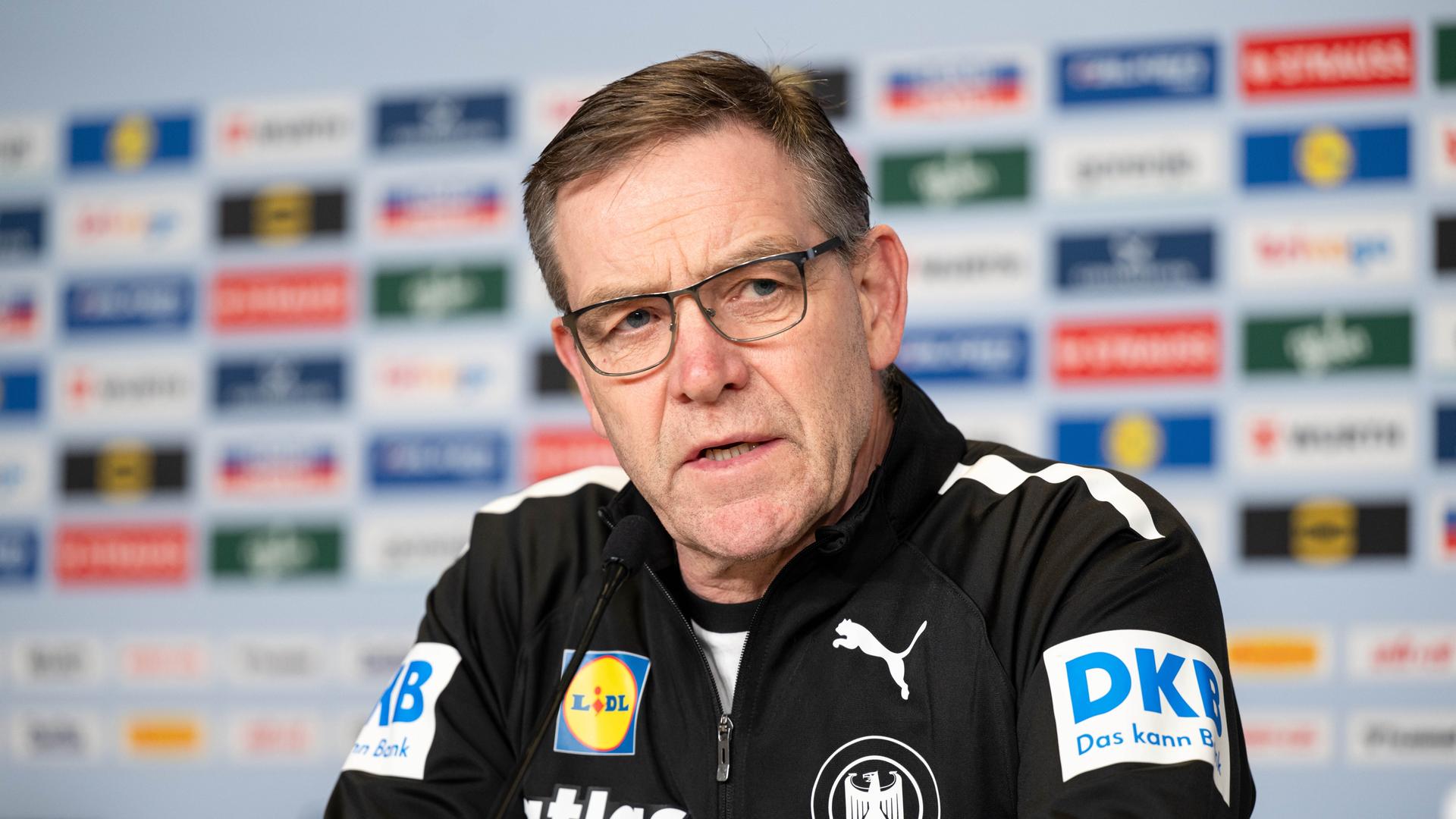 Alfred Gislason spricht während einer Pressekonferenz vor dem Halbfinale Deutschland gegen Kroatien. Er trägt einen schwarzen Pullover mit mehreren bunten Logos.