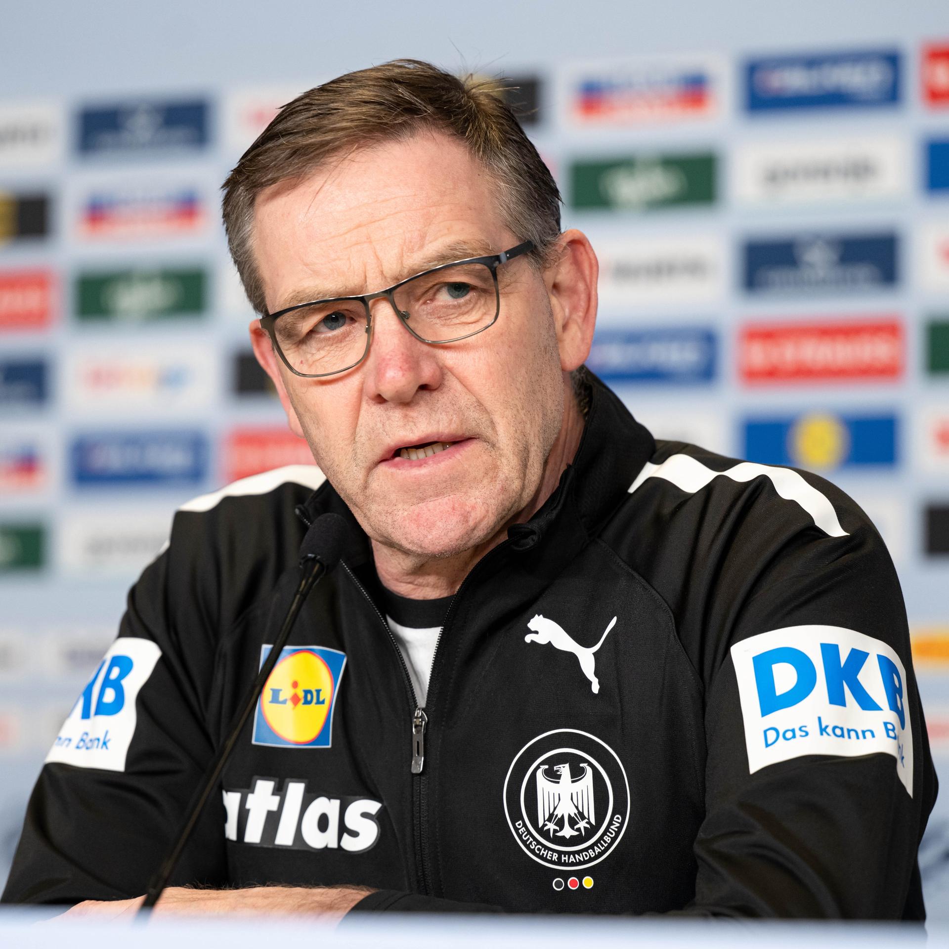 Alfred Gislason spricht während einer Pressekonferenz vor dem Halbfinale Deutschland gegen Kroatien. Er trägt einen schwarzen Pullover mit mehreren bunten Logos.