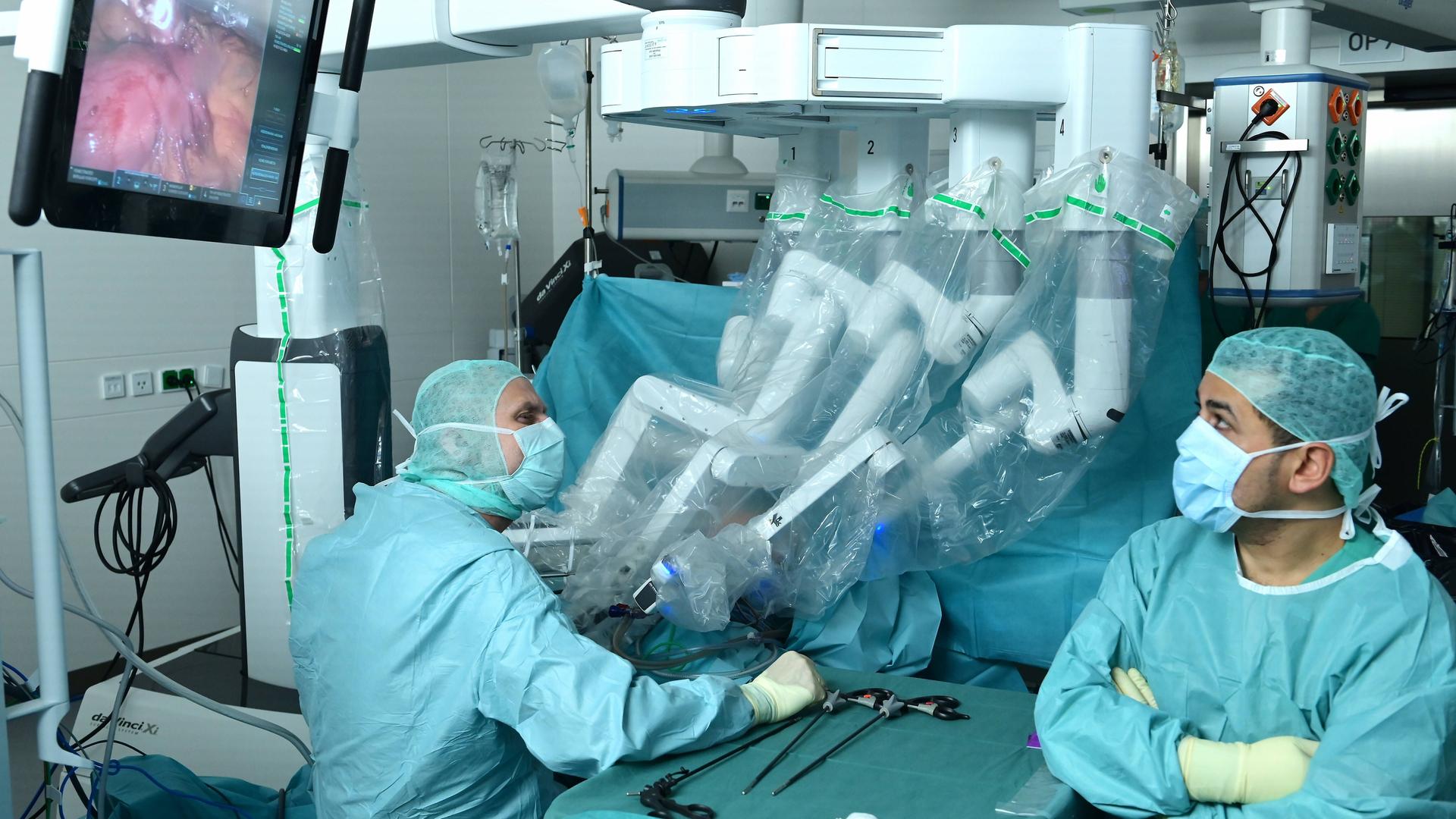 Zwei Ärzte sitzen im OP mit einem Roboter, der mit Armen bei der OP assistiert. 