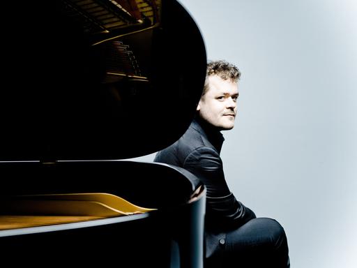 Der Pianist Benjamin Grosvenor sitzt am hinteren Ende eines aufgeklappten Flügels und schaut an der Rundung des Instrumentes vorbei.