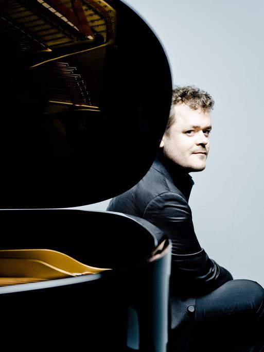 Der Pianist Benjamin Grosvenor sitzt am hinteren Ende eines aufgeklappten Flügels und schaut an der Rundung des Instrumentes vorbei.