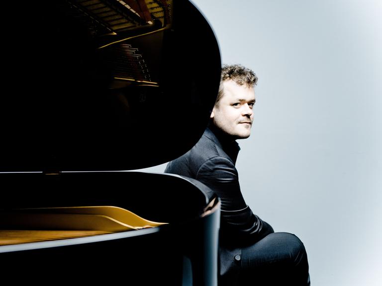Der Pianist Benjamin Grosvenor sitzt am hinteren Ende eines aufgeklappten Flügels und schaut an der Rundung des Instrumentes vorbei.