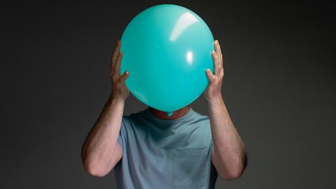 Porträt eines Mannes, der einen hellblauen Luftballon vor sein Gesicht hält.