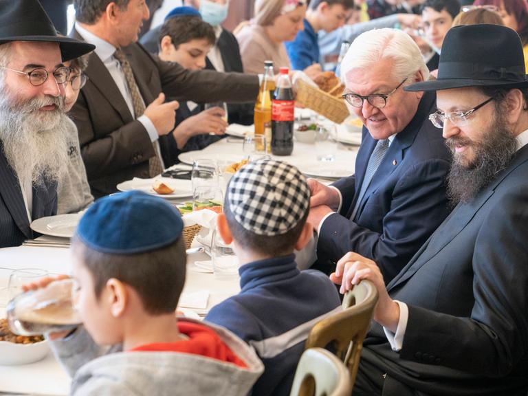 Frank-Walter Steinmeier sitzt mit orthodox gekleideten Juden an einem Tisch beim Essen und spricht mit aus der Ukraine geflüchteten Kindern.