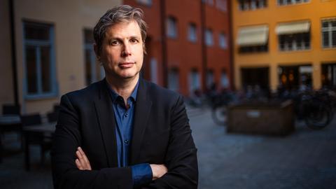 Daniel Kehlmann steht mit verschränkten Armen in einem Innenhof und blickt ernst in die Kamera.