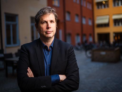 Daniel Kehlmann steht mit verschränkten Armen in einem Innenhof und blickt ernst in die Kamera.