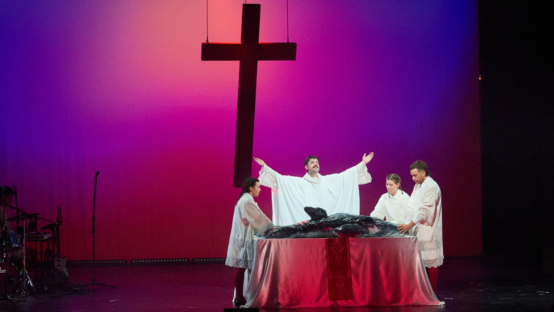 Vier Schauspieler stehen in religiös anmutenden Gewändern um einen Tisch, der wie ein Altar aussieht. Auf dem Tisch liegt ein aufblasbarer Wal. Im Hintergrund schwebt ein Kreuz. Vier Schauspieler stehen in religiös anmutenden Gewändern um einen Tisch, der wie ein Altar aussieht. Auf dem Tisch liegt ein aufblasbarer Wal. Im Hintergrund schwebt ein Kreuz.