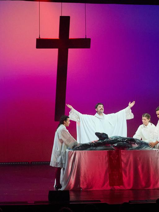 Vier Schauspieler stehen in religiös anmutenden Gewändern um einen Tisch, der wie ein Altar aussieht. Auf dem Tisch liegt ein aufblasbarer Wal. Im Hintergrund schwebt ein Kreuz. 