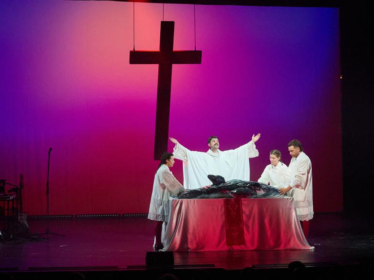 Vier Schauspieler stehen in religiös anmutenden Gewändern um einen Tisch, der wie ein Altar aussieht. Auf dem Tisch liegt ein aufblasbarer Wal. Im Hintergrund schwebt ein Kreuz. 