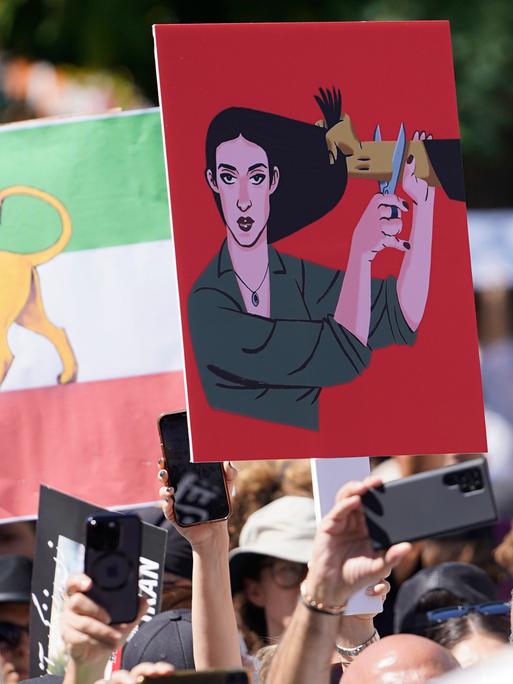 Auf einer Demonstration sieht man ein Plakat. Auf dem Plakat ist eine Frau zu sehen, der ein Mann ins Haar greift. Die Frau hält eine Haarschere an den Arm des Mannes.