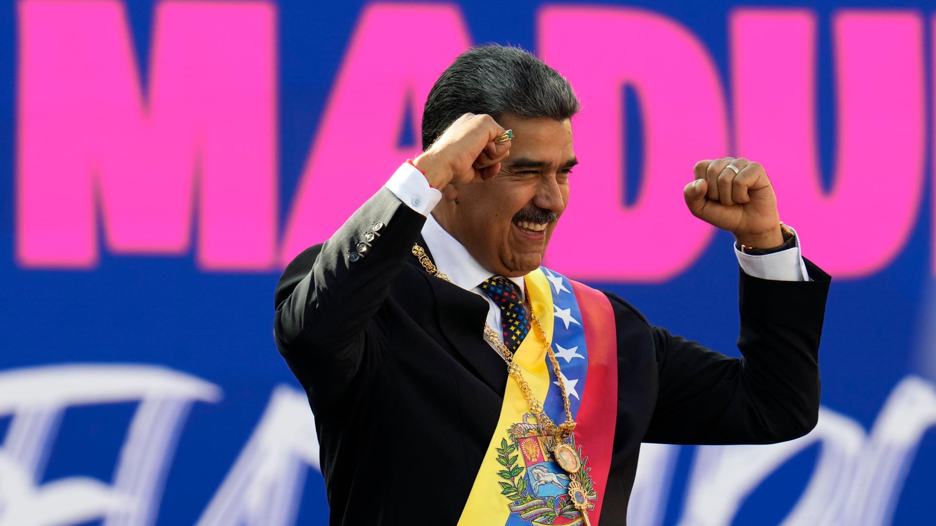 Nicolas Maduro ballt die Fäuste in einer Sieger-Pose und lacht. Dahinter ein pinker Schriftzug auf blauem Grund mit seinem Namen. Im Bild sind nur die Buchstaben MADU