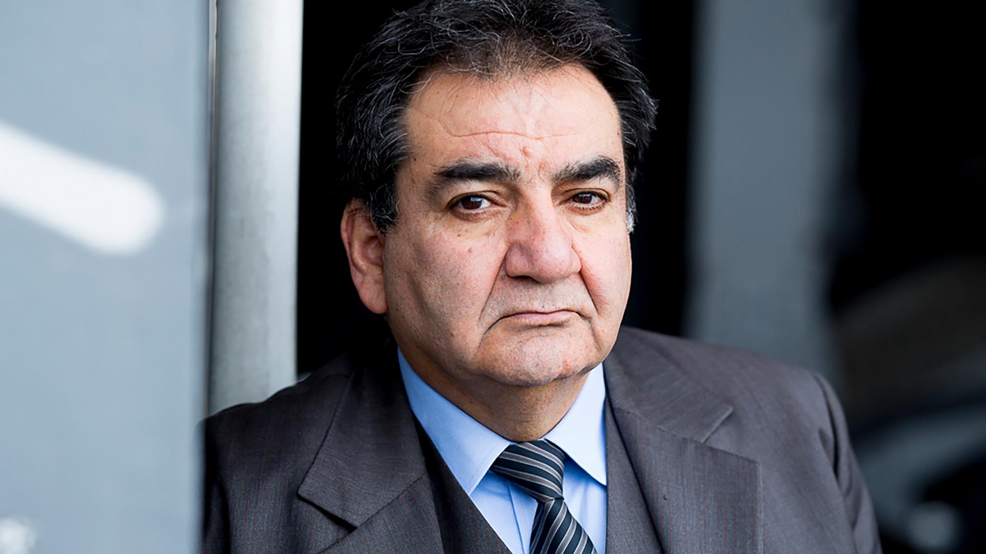 Mohammad-Ali Behboudi trägt einen grauen Anzug und Krawatte, lehnt an einer Wand und schaut ernst in die Kamera. Mohammad-Ali Behboudi trägt einen grauen Anzug und Krawatte, lehnt an einer Wand und schaut ernst in die Kamera.