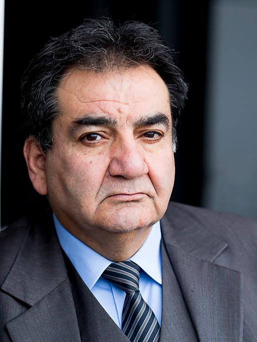 Mohammad-Ali Behboudi trägt einen grauen Anzug und Krawatte, lehnt an einer Wand und schaut ernst in die Kamera.