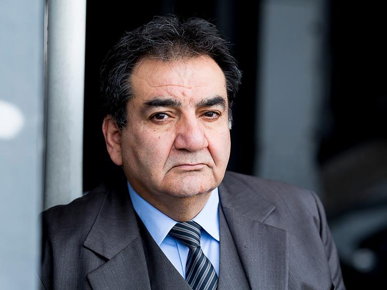 Mohammad-Ali Behboudi trägt einen grauen Anzug und Krawatte, lehnt an einer Wand und schaut ernst in die Kamera.