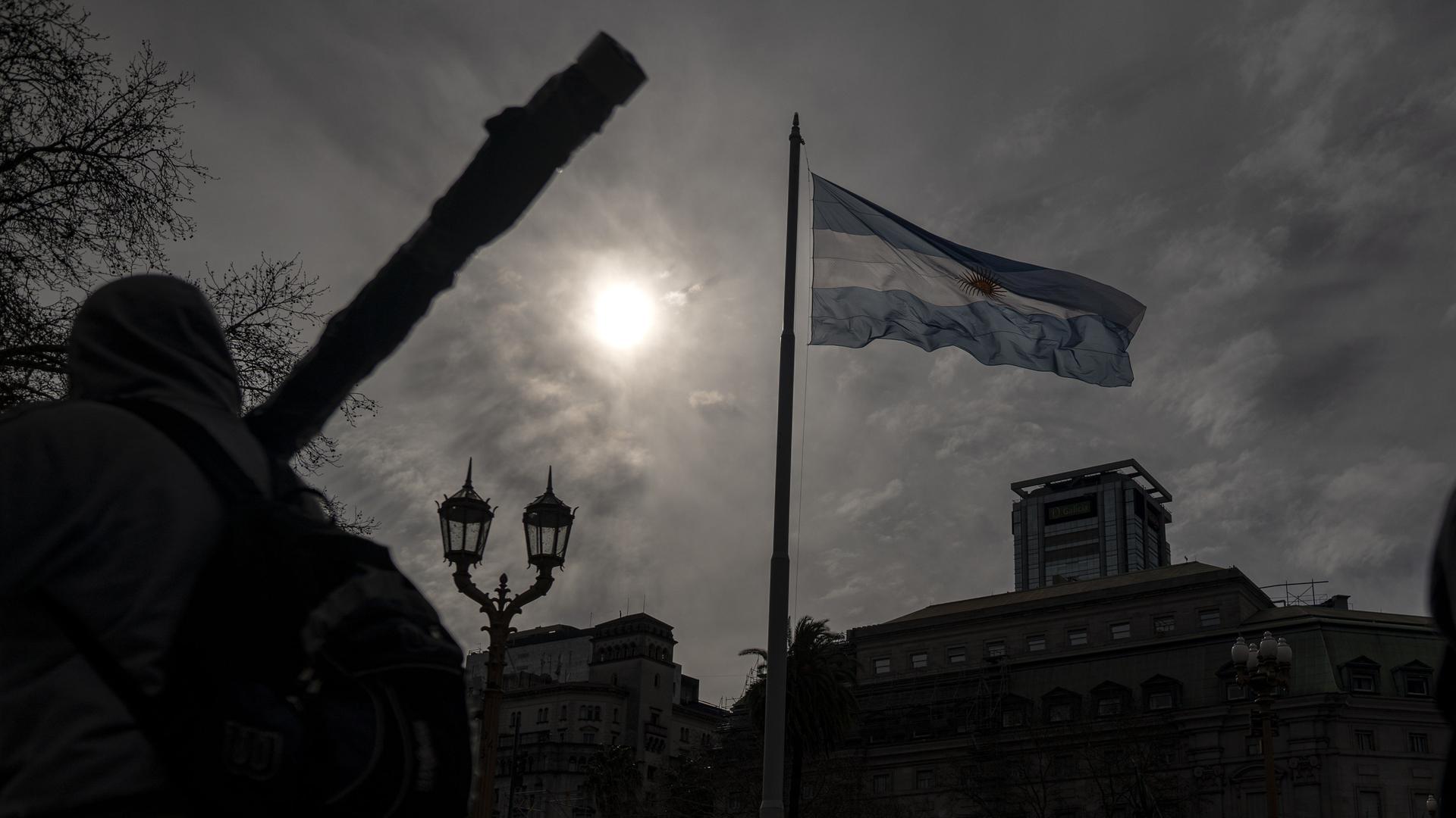 Die Flagge Argentiniens weht über einer Protestaktion in Buenos Aires, während der Himmel so trüb ist, dass die Sonne nur ein fahles Licht auf die Straße strahlt.