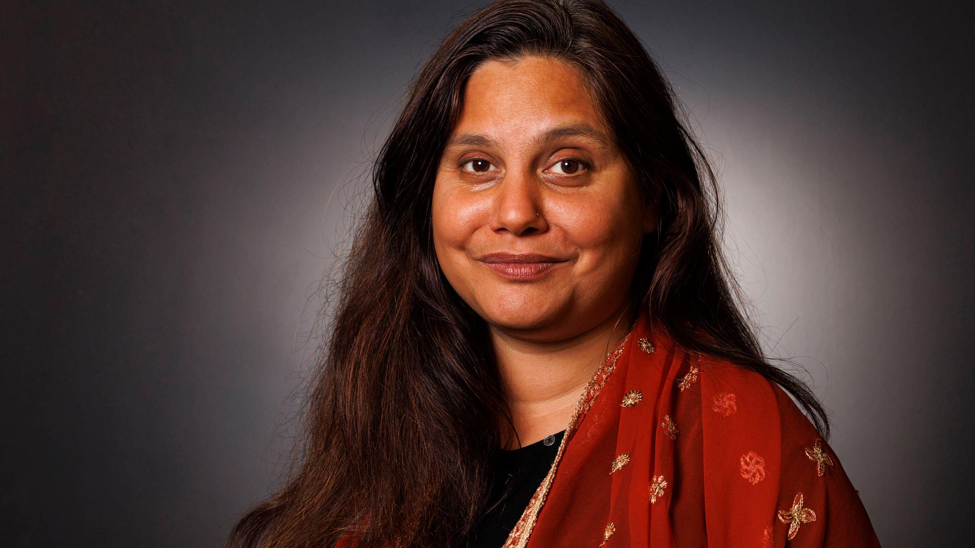 Porträtaufnahme von Mithu Sanyal vor grauer Wand. Sie lächelt in die Kamera und trägt ein farbenfrohes Kleid Porträtaufnahme von Mithu Sanyal vor grauer Wand. Sie lächelt in die Kamera und trägt ein farbenfrohes Kleid