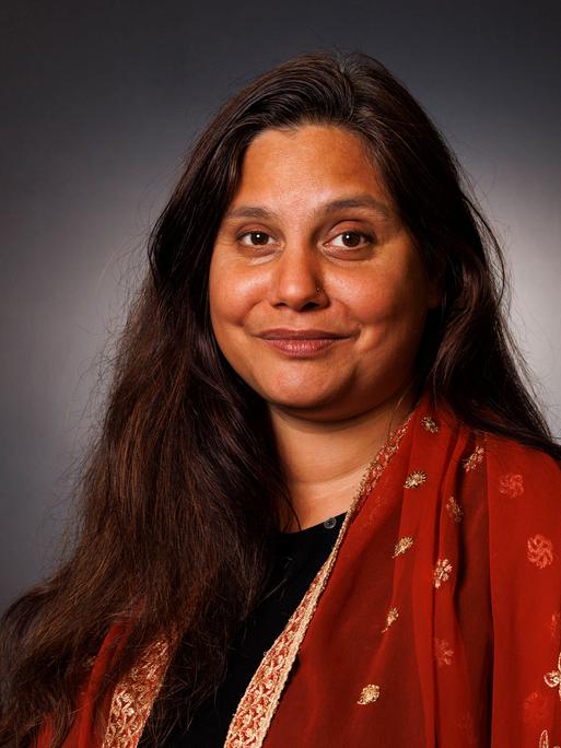 Porträtaufnahme von Mithu Sanyal vor grauer Wand. Sie lächelt in die Kamera und trägt ein farbenfrohes Kleid