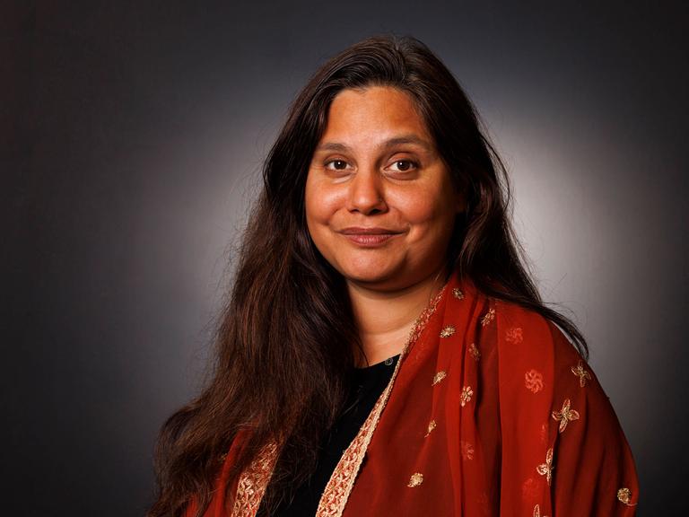 Porträtaufnahme von Mithu Sanyal vor grauer Wand. Sie lächelt in die Kamera und trägt ein farbenfrohes Kleid