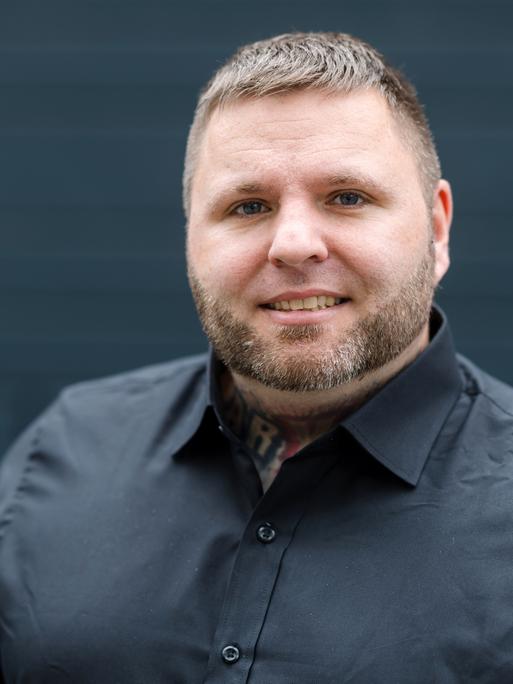 Tommy Frenck steht in schwarzem Hemd vor schwarzem Hintergrund und guckt in die Kamera.