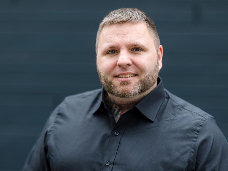 Tommy Frenck steht in schwarzem Hemd vor schwarzem Hintergrund und guckt in die Kamera.