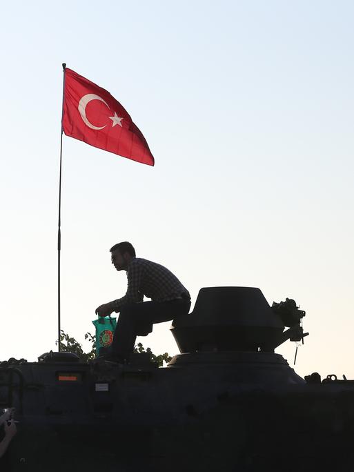 Die Silhouette eines Mannes auf einem gepanzerten Fahrzeug zeichnet sich gegen den Morgenhimmel ab. Auf dem Fahrzeug ist die türkische Nationalflagge gehisst. Die Silhouette eines Mannes auf einem gepanzerten Fahrzeug zeichnet sich gegen den Morgenhimmel ab. Auf dem Fahrzeug ist die türkische Nationalflagge gehisst.