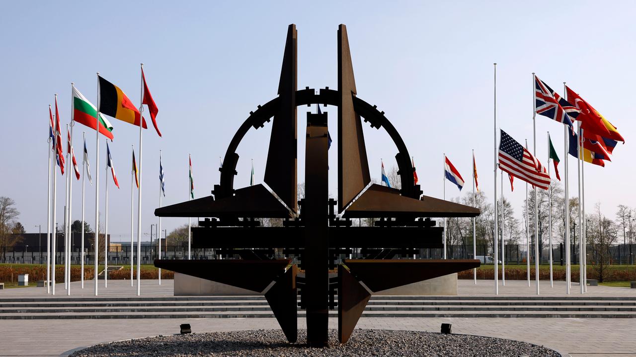 Blick auf die NATO-Sternenskulptur am Sitz der NATO in Brüssel, im Hintergrund wehen die Flaggen der Mitgliedstaaten an Fahnenmasten.