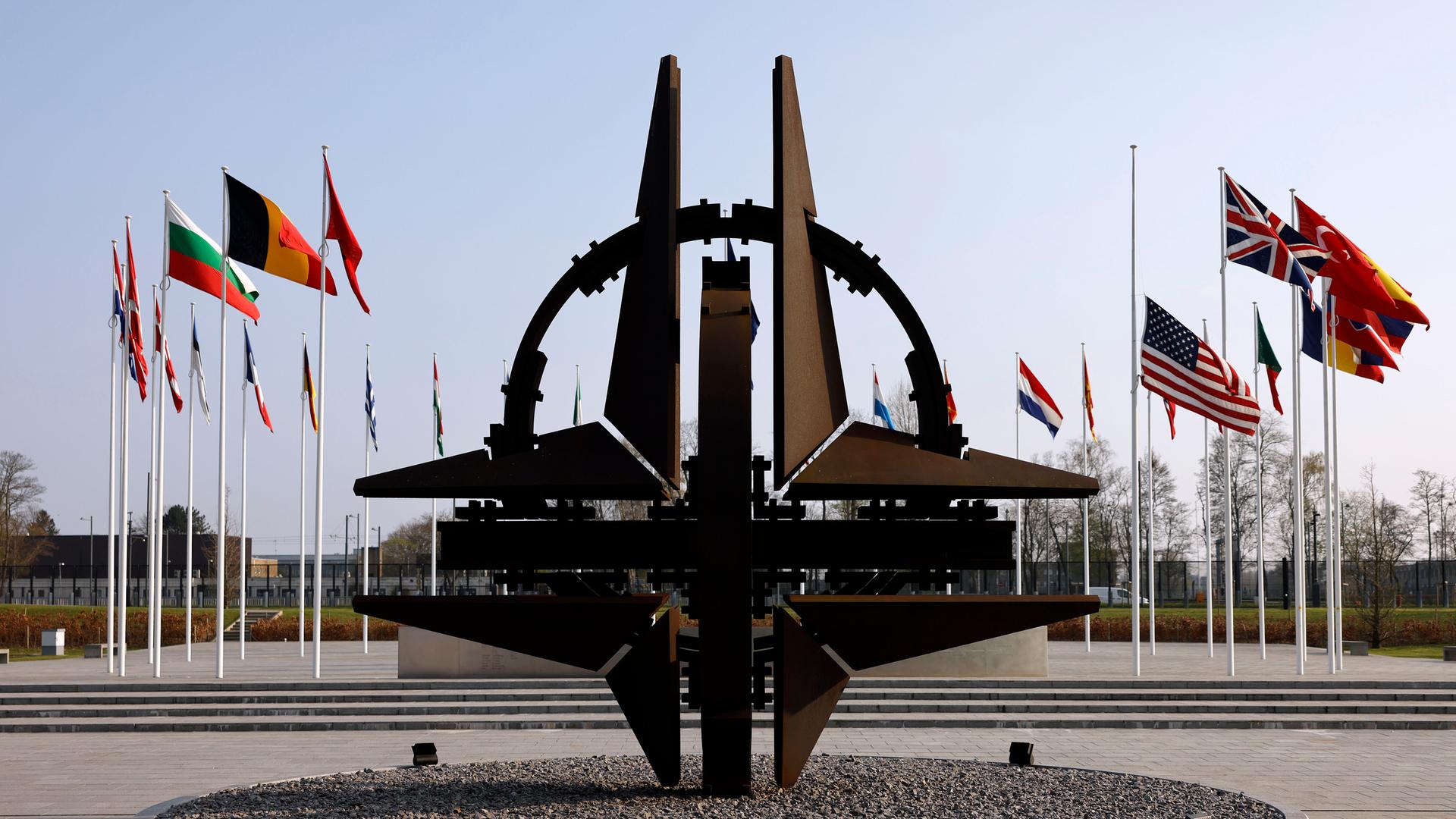 Blick auf die NATO-Sternenskulptur am Sitz der NATO in Brüssel, im Hintergrund wehen die Flaggen der Mitgliedstaaten an Fahnenmasten.