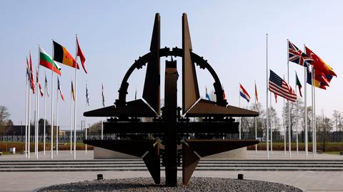 Blick auf die NATO-Sternenskulptur am Sitz der NATO in Brüssel, im Hintergrund wehen die Flaggen der Mitgliedstaaten an Fahnenmasten.