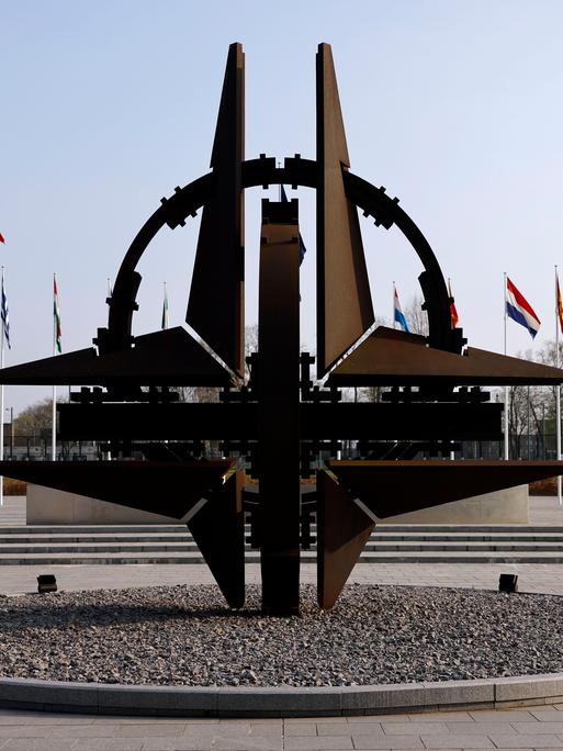 Blick auf die NATO-Sternenskulptur am Sitz der NATO in Brüssel, im Hintergrund wehen die Flaggen der Mitgliedstaaten an Fahnenmasten.