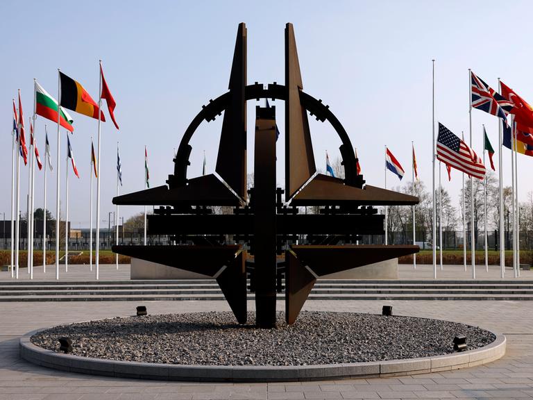 Blick auf die NATO-Sternenskulptur am Sitz der NATO in Brüssel, im Hintergrund wehen die Flaggen der Mitgliedstaaten an Fahnenmasten.