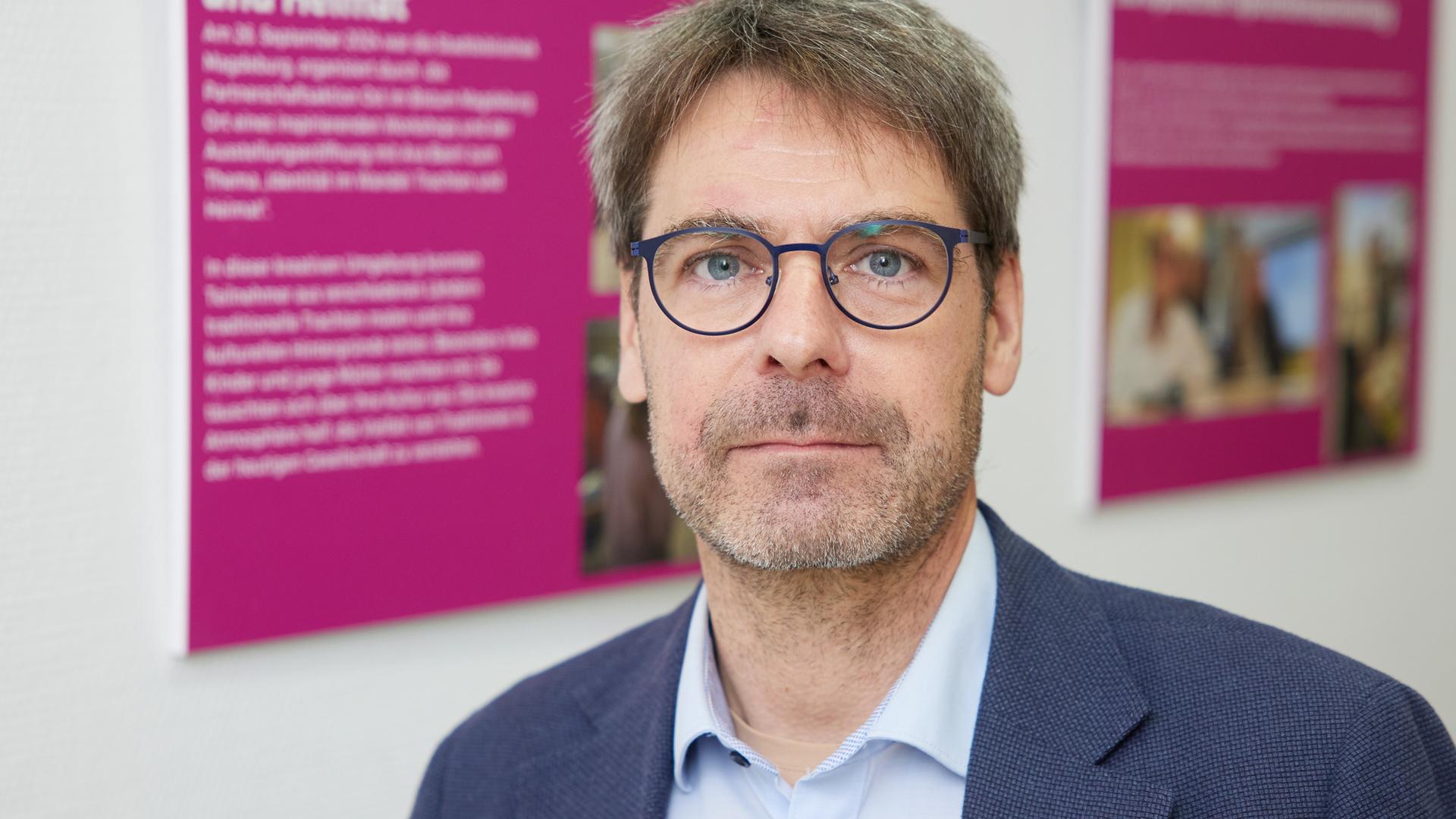 Porträt eines Mannes mit kurzen, braunen Haaren und Brille in blauem Sakko.