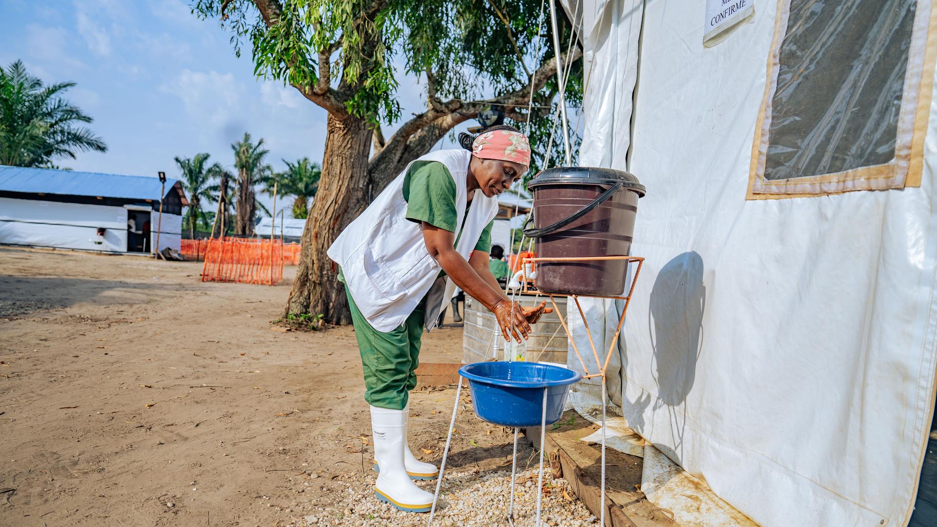 Eine Krankenschwester in Schutzkleidung wäscht ihre Hände an einem Wassercontainer neben improvisierten Krankenzelten. Eine Krankenschwester in Schutzkleidung wäscht ihre Hände an einem Wassercontainer neben improvisierten Krankenzelten.