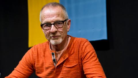 Gerbrand Bakker sitzt im orangen Pulli an einem Tisch und blickt ernst in die Kamera